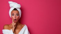 Young woman in white bath towel and turban making shush gesture with finger on lips against pink background, expressing surprise with wide eyes and open mouth.