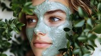 Young woman wearing green clay face mask surrounded by fresh green leaves, representing natural skincare and beauty wellness routines in spa setting.