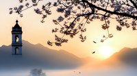 Church bell tower silhouetted against mountain landscape at sunset with cherry blossom branches and flying birds creating peaceful spiritual atmosphere.