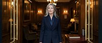 Professional blonde businesswoman in navy suit standing confidently in luxury hotel lobby with warm lighting and elegant interior design for corporate use.