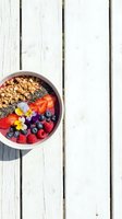 Colorful acai bowl topped with granola, fresh berries, chia seeds and edible flowers on white wooden surface, ideal for healthy eating and wellness content.