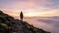 Hiker standing on mountain ridge at sunrise above clouds, wearing backpack and outdoor gear, enjoying scenic landscape view during golden hour adventure.
