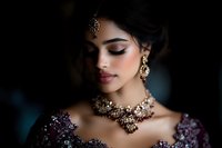 Young South Asian woman wearing traditional bridal jewelry and purple embroidered dress, with elegant makeup and closed eyes against dark background.