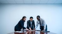 Diverse business team of three professionals collaborating over documents at conference table in modern office meeting room for strategic planning.