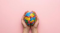 Hands holding colorful brain model with vibrant sections on pink background representing mental health awareness and cognitive development concepts.