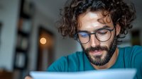 Young Mediterranean man with curly hair and glasses working on laptop at home office, focused on digital screen in evening. Professional freelancer in teal shirt.