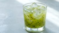 Refreshing kiwi cocktail in a glass with ice cubes on a light gray surface, featuring green juice and black seeds for summer drink menus.