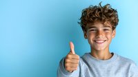 Young Hispanic teen boy with curly hair and bright smile showing thumbs up gesture against blue background, wearing casual grey t-shirt. Positive youth expression.