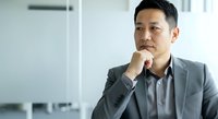 Asian male professional in business casual attire sitting thoughtfully with hand to chin, contemplating ideas in modern office environment for corporate, leadership, and business concept use.