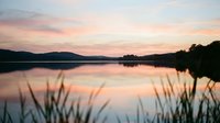 Serene lake at sunset with calm water reflecting pink and orange sky, silhouetted mountains and trees, marsh grasses in foreground. Perfect for nature, travel, and wellness projects.