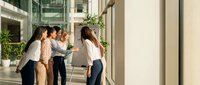 Diverse business team of young professional women collaborating in modern office corridor with natural lighting and plants.