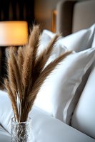 Dried pampas grass in crystal vase against white bedding and warm lamp light creates cozy bedroom atmosphere. Modern minimalist home decor styling.