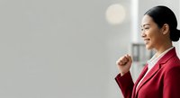 Asian woman wearing red blazer and glasses, smiling with fist raised in confident gesture against blurred office background, representing success, motivation and professional achievement.