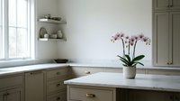 Young woman with light skin tone, wearing casual home attire, arranging fresh pink orchids in a minimalist kitchen with sage green cabinetry and white marble countertops for interior design and lifestyle content.