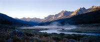 Serene mountain lake at dawn with misty waters, forested slopes, and golden sunlit peaks reflected in calm alpine landscape.