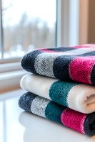 Stack of folded knitted sweaters in colorful geometric patterns near window. Cozy winter clothing collection with pink, teal, navy and cream colors for seasonal wardrobe.