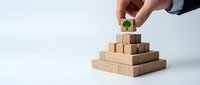 Hand placing wooden block with green tree symbol on pyramid of natural wood cubes representing environmental growth and sustainable business development.