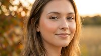Young Caucasian woman with long brown hair smiling warmly at camera during golden hour sunset with soft bokeh background for lifestyle content.
