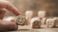 Hand picking a wooden cube with a smiley face among sad face blocks on a table, symbolizing positive choice and happiness.