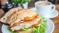Fresh grilled chicken sandwich with lettuce, tomato and melted cheese on artisanal bread, served with hot cappuccino on wooden table, close up view.
