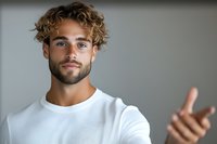 Young Caucasian male with curly blonde hair and beard wearing white t-shirt gesturing with hand against gray background, expressing confidence and positive attitude.