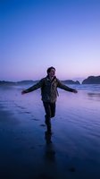 Man in casual jacket running on wet beach at twilight with arms outstretched, enjoying freedom and nature during purple sunset hour with mountains in background.