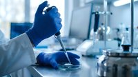 Scientist in laboratory conducting research with pipette and petri dish, wearing blue gloves and white coat in sterile environment.
