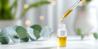 Dropper adding golden essential oil to small glass bottle, with soft-focus eucalyptus leaves and fairy lights creating spa-like atmosphere for aromatherapy or skincare routine.