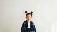 Young Asian girl with black hair styled in two buns, wearing a dark denim jacket over a white shirt, smiling at camera against neutral background for portrait and lifestyle use.