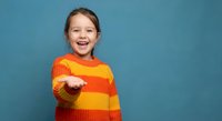 Happy young Caucasian girl in colorful striped sweater pointing forward with joyful expression on blue background for children advertising campaigns.