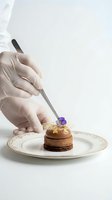 Professional chef in white gloves using tweezers to place purple flower garnish on elegant chocolate dessert with gold leaf decoration on fine dining plate.