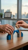 Two hands stacking wooden blocks on office desk with city skyline view through window, representing teamwork, business growth, and corporate collaboration in modern workplace setting.
