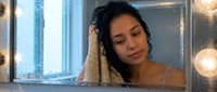 Young woman with dark curly hair and medium skin tone looking at mirror with warm lighting, suitable for beauty, skincare, wellness and personal care marketing campaigns.