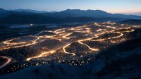 Aerial view of glowing city lights creating organic patterns across mountain valley at dusk, illuminated roads weaving through residential areas against dark peaks.