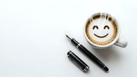 Coffee cup with smiley face foam art next to black pens on white background. Morning motivation and positive workspace concept for office productivity.
