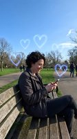 Young woman with curly hair texting on phone in park with floating heart symbols representing online dating and digital romance connections.
