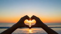 Hands forming heart shape silhouette against golden sunset over ocean waves. Romantic gesture symbolizing love, connection and peaceful moments by the sea.