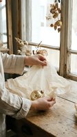 Woman hands carefully wrapping golden Christmas ornament in white tissue paper on rustic wooden table by window with dried flowers for holiday storage.