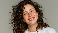 Young woman with curly brown hair and bright smile wearing white top against neutral background, conveying happiness and confidence.