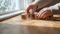 Hands arranging wooden blocks with renewable energy icons including solar panel, wind turbine and leaf symbols on wooden surface near window.
