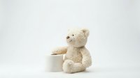 Soft beige teddy bear sitting beside white ceramic bowl on clean white background, isolated for nursery decor, baby shower invitations, childcare branding.