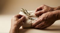 Elderly hands carefully wrapping white gift box with natural twine and dried plant decoration on warm beige background for special occasions.