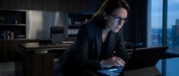 Mature woman with glasses wearing black business suit working on laptop at modern office desk with city skyline view at night, professional corporate environment.
