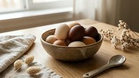 Fresh farm eggs in rustic ceramic bowl on wooden cutting board with natural morning light streaming through kitchen window for cooking and baking.