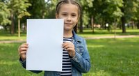 Young girl holding blank white paper sheet in park for mockup design projects and educational presentations.
