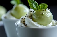Green grape dessert with whipped cream and black sesame seeds garnished with fresh mint leaf in white ceramic bowl on dark background, macro food photography.
