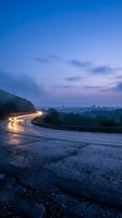 Winding mountain road with car light trails at twilight creating dynamic motion blur against purple sky and distant city lights for transportation concepts.