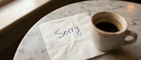 Handwritten sorry note on a napkin beside a black coffee cup on a marble table, conveying apology, regret, and reconciliation in a cafe setting.