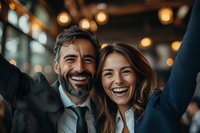Joyful mature business couple celebrating success with arms raised, genuine emotional moment captured in warm ambient lighting with bokeh background.