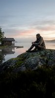Solitary man in outdoor gear sits on mossy rock by misty lake at dawn, wooden cabin and boat visible in serene Nordic wilderness landscape.